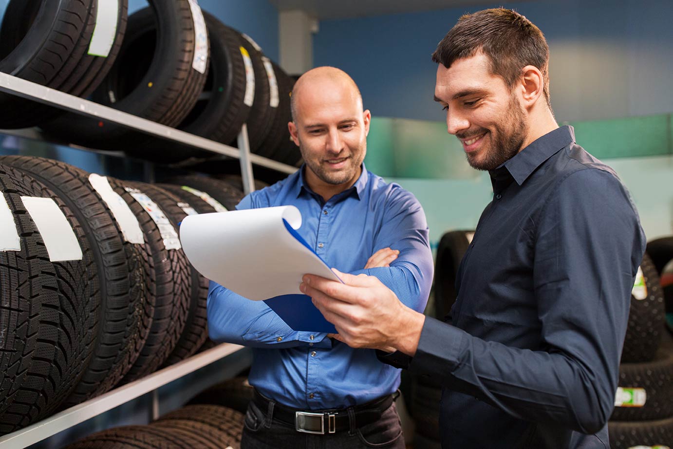 two men looking at a piece of paper on a clipboard and smiling while standing next to a wall rack of tires